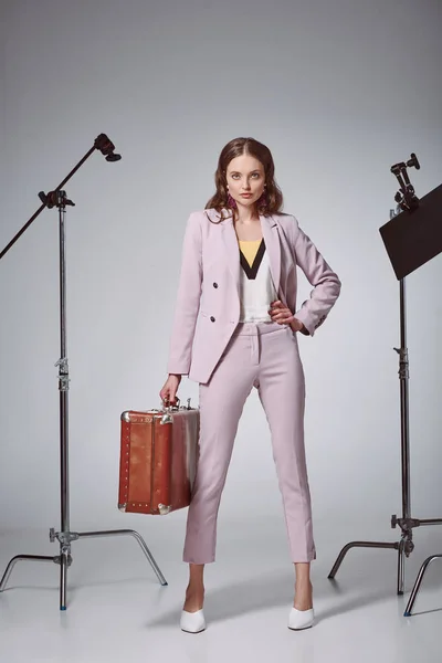 Belle femme à la mode avec valise posant à la main sur la taille et regardant la caméra en studio d'enregistrement — Photo de stock