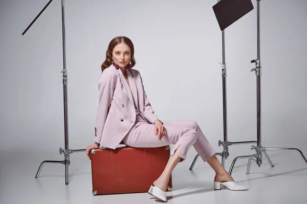 Fashionable woman in pink suit sitting on suitcase and looking at camera in recording studio — Stock Photo