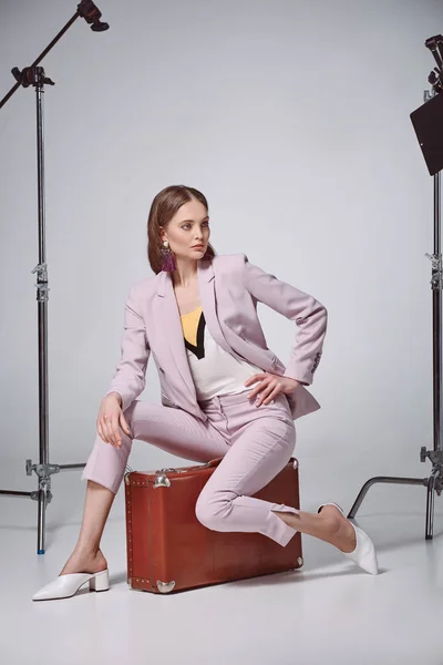 Femme à la mode en costume rose assis sur la valise et regardant loin dans le studio d'enregistrement — Photo de stock