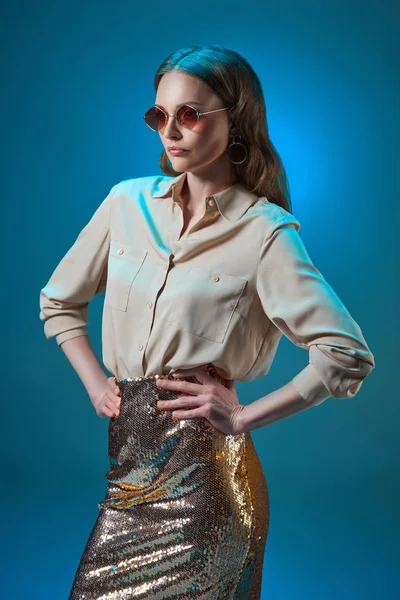 Beautiful woman in fashionable golden glittering skirt and sunglasses posing with hands on waist isolated on blue — Stock Photo