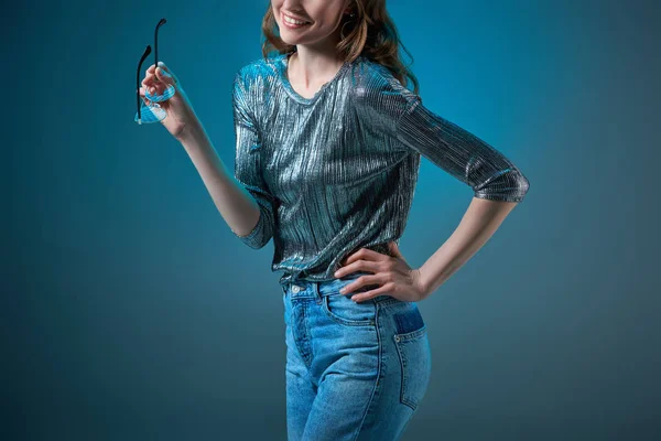 Recortado disparo de mujer sonriente sosteniendo gafas y posando con la mano en la cintura aislado en azul - foto de stock