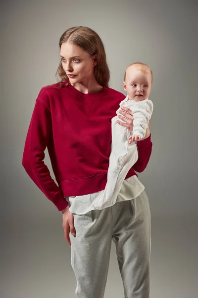Hermosa madre elegante sosteniendo adorable hija lactante y mirando hacia otro lado aislado en gris - foto de stock