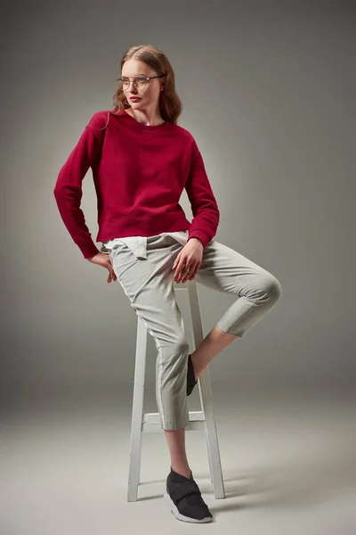 Full length view of beautiful woman in stylish outfit and spectacles sitting on stool and looking away on grey — Stock Photo