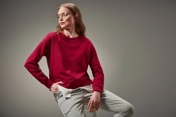 Hermosa mujer en suéter rojo sentado en el taburete y mirando hacia otro lado aislado en gris - foto de stock