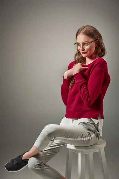 Schöne lächelnde Frau mit Brille, die auf einem Schemel sitzt und isoliert von grau wegschaut — Stockfoto