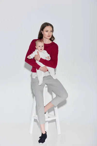 Full length view of beautiful stylish woman holding adorable baby girl and sitting stool on grey — Stock Photo