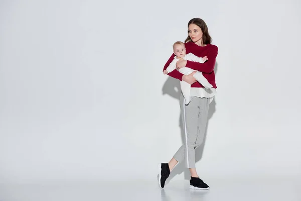 Vista completa de la hermosa madre joven en traje elegante llevando adorable hija bebé en gris - foto de stock