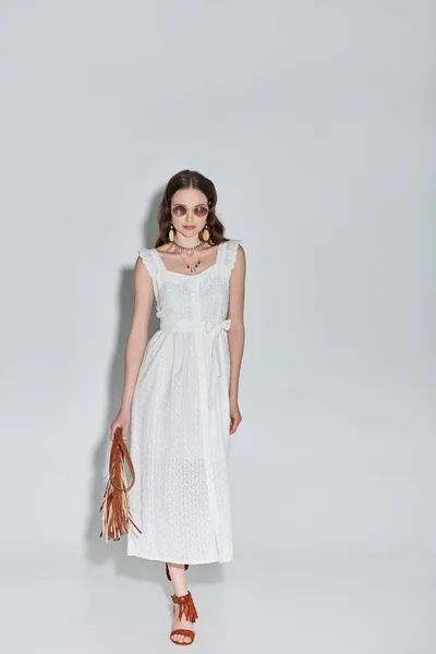 In voller Länge Ansicht der schönen Frau in stilvollem weißen Kleid und Sonnenbrille mit Handtasche und Fuß auf grau — Stockfoto
