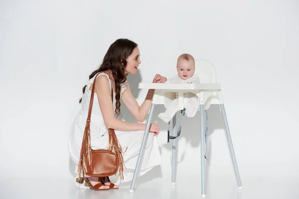 Mulher sorridente elegante olhando para a filha bebê adorável sentado em cadeira alta no cinza — Fotografia de Stock
