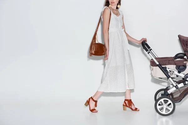 Plan recadré de jeune femme en robe blanche à la mode marchant avec chariot bébé sur gris — Photo de stock