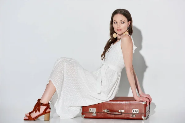 Beautiful woman in stylish white dress sitting on retro suitcase and looking away on grey — Stock Photo