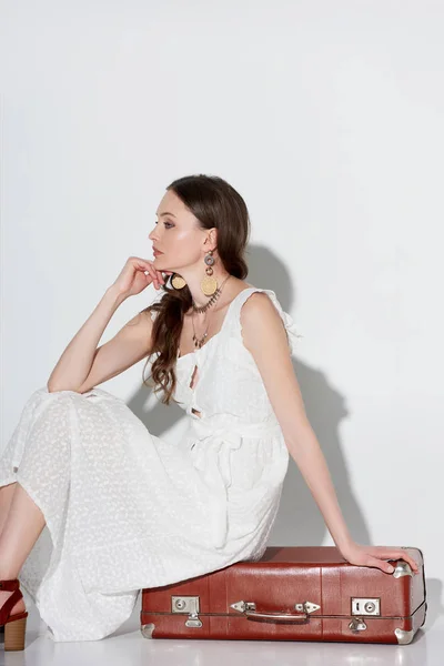 Beautiful pensive young woman in stylish white dress sitting on retro suitcase and looking away on grey — Stock Photo