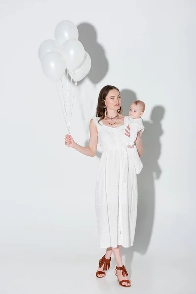 Belle femme en robe blanche élégante portant adorable bébé fille et tenant tas de ballons sur gris — Photo de stock