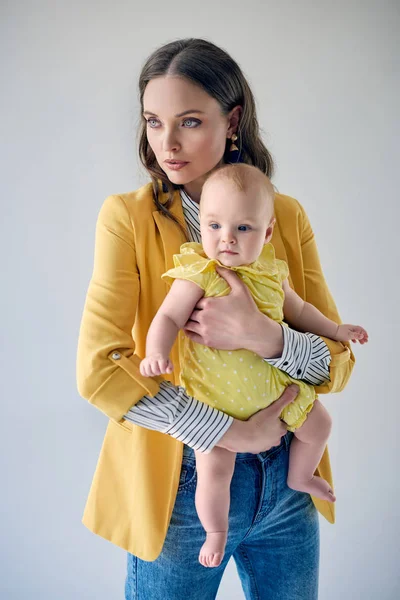 Young mother in stylish outfit carrying adorable infant daughter and looking away isolated on grey — Stock Photo