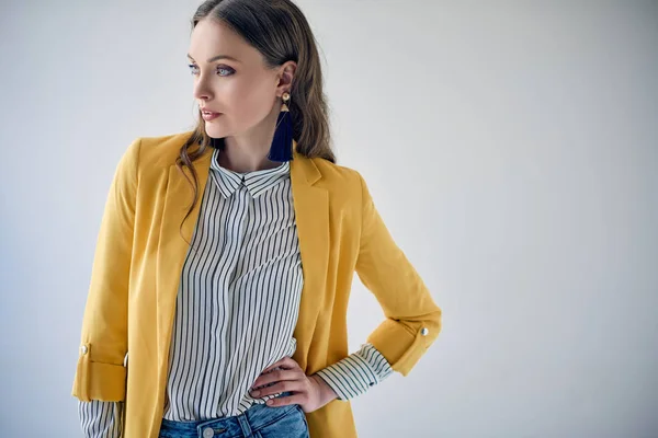 Schöne junge Frau in gelber Jacke und gestreiftem Hemd posiert mit der Hand auf der Taille und schaut isoliert auf grau weg — Stockfoto