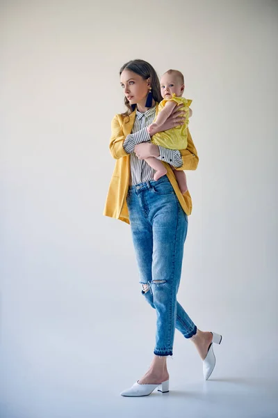 Vue pleine longueur de la jeune mère élégante portant adorable fille bébé et marchant sur gris — Photo de stock