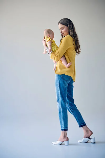 Vista lateral de hermosa madre joven con estilo llevando niño adorable en gris - foto de stock