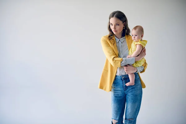 Madre joven de moda llevando a su adorable hija lactante y mirando hacia otro lado aislado en gris - foto de stock