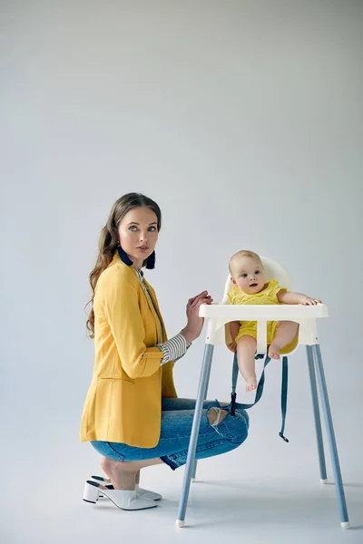 Красивая модная мать, приседающая рядом с младенческой дочерью, сидящей в высоком кресле на сером — стоковое фото