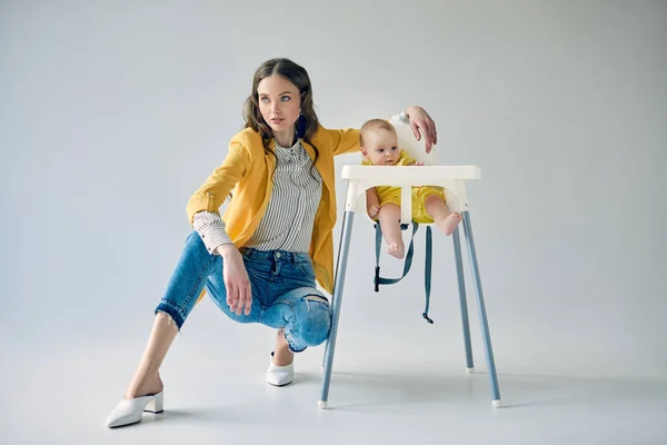 Beautiful stylish mother crouching near infant daughter sitting in high chair on grey — Stock Photo