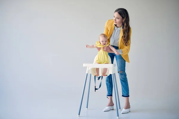 Schöne stilvolle Mutter setzt Baby-Mädchen im Hochstuhl auf grau — Stockfoto