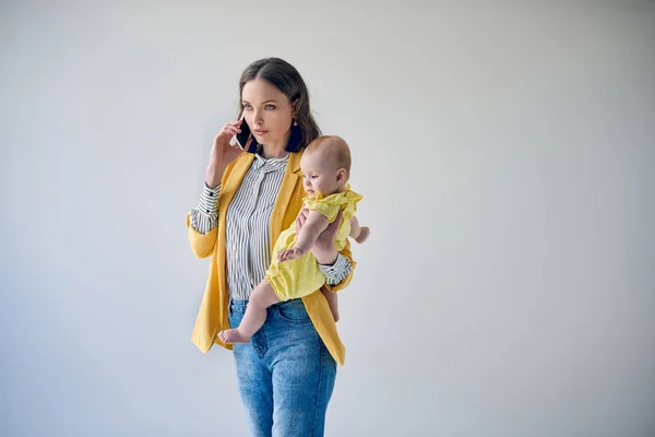 Belle mère élégante portant adorable petite fille et parlant par smartphone isolé sur gris — Photo de stock