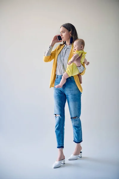 Madre elegante llevando adorable hija lactante y hablando por teléfono inteligente en gris - foto de stock