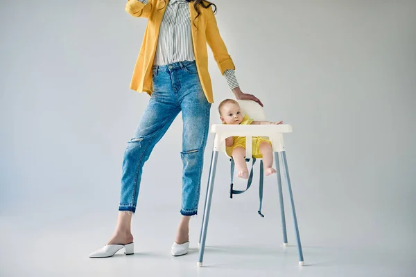 Plan recadré de la mère élégante debout près de bébé fille assis dans une chaise haute sur gris — Photo de stock