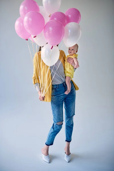 Vista completa de la mujer elegante llevando adorable bebé niña y sosteniendo globos en gris - foto de stock