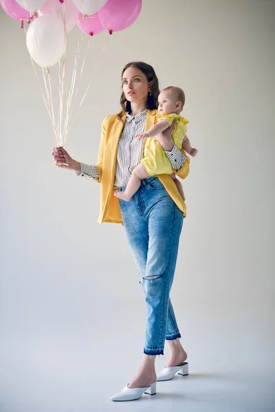 Vista basso angolo di donna elegante che trasporta adorabile bambina e tenendo mazzo di palloncini su grigio — Foto stock