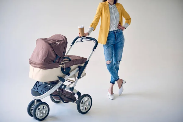 Tiro recortado de mulher elegante segurando copo de café descartável e de pé com carrinho de bebê em cinza — Fotografia de Stock