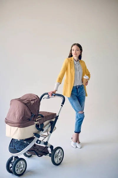 Stylische junge Frau hält Einweg-Kaffeetasse, während sie mit Kinderwagen steht und in die Kamera auf grau blickt — Stockfoto