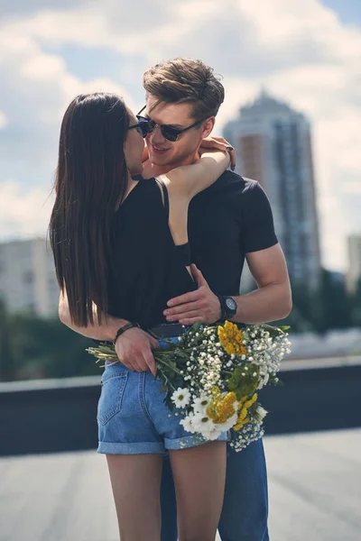 Sexy multiethnic couple embracing and holding bouquet of flowers on roof — Stock Photo