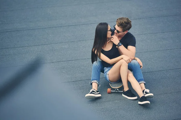 Schönes multiethnisches Paar, das sich umarmt und auf dem Skateboard sitzt — Stockfoto