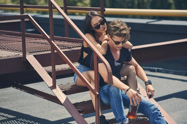 Многонациональная пара с бутылкой виски, сидящей на лестнице на крыше — стоковое фото
