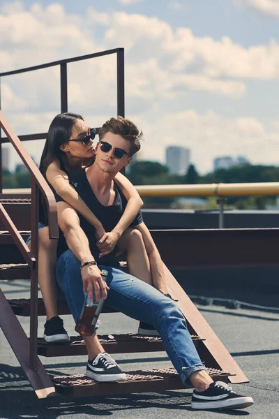 Young multiethnic couple with bottle of whiskey sitting on stairs on roof — Stock Photo
