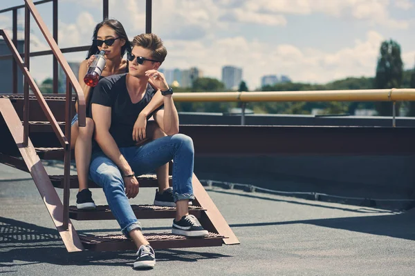 Pareja interracial con botella de ron sentado en las escaleras en el techo - foto de stock