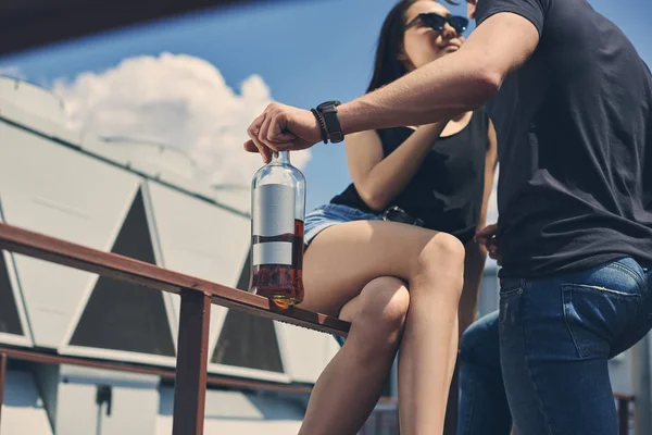 Hot multicultural couple flirting and holding bottle of rum on roof — Stock Photo