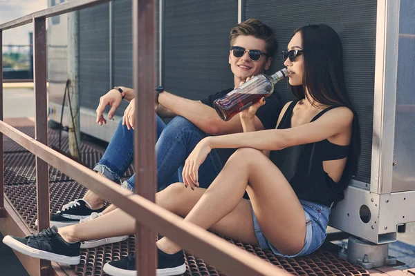 Young interracial couple drinking whiskey from bottle on roof — Stock Photo