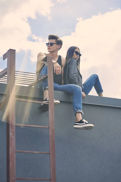 Stylish multiethnic couple in sunglasses sitting on roof with sunlight — Stock Photo