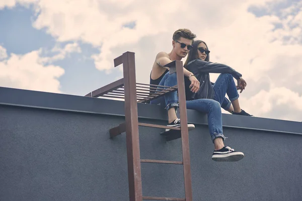 Stylish multicultural couple in sunglasses sitting on roof with ladder — Stock Photo
