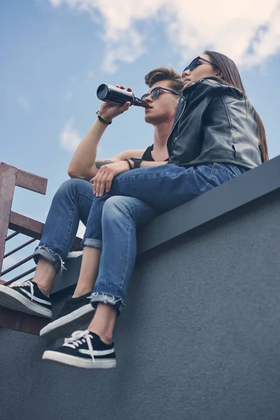 Bottom view of stylish interracial couple in sunglasses sitting on roof, man drinking beer — Stock Photo