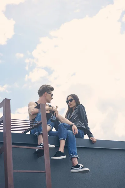 Elegante pareja joven interracial en gafas de sol sentado en el techo con escalera - foto de stock