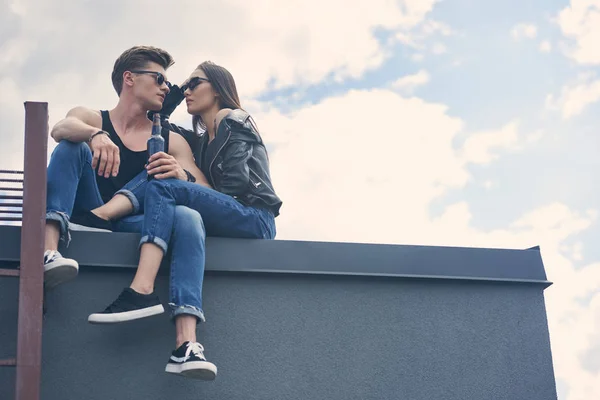 Schönes multikulturelles Paar mit Sonnenbrille auf dem Dach sitzend mit einer Flasche Bier — Stockfoto