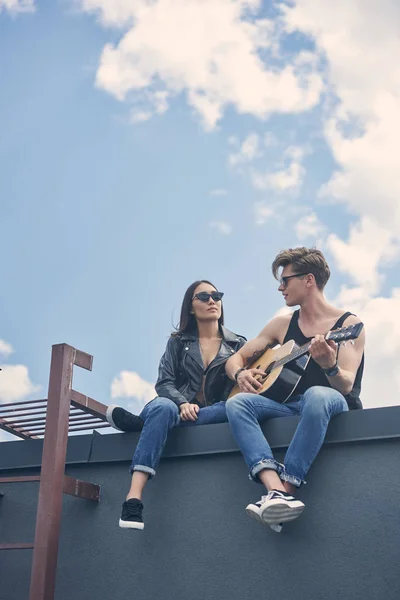 Guapo guitarrista jugando para su asiático novia mientras pareja sentado en techo - foto de stock