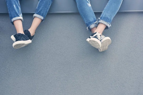 Vista parcial de un par de piernas en jeans y zapatillas de deporte sentadas en la pared gris - foto de stock