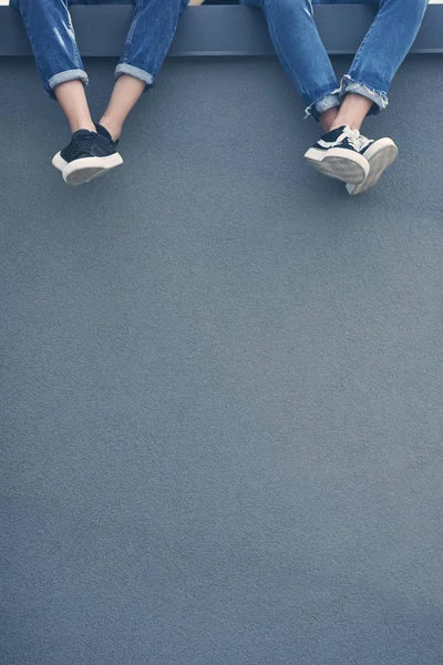 Vista recortada de pareja en jeans y zapatillas de deporte sentados en la pared gris - foto de stock