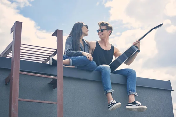 Feliz pareja multicultural sentado en el techo con guitarra acústica - foto de stock