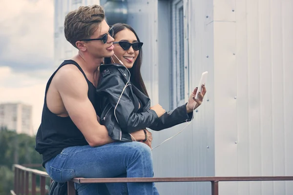 Улыбающаяся мультикультурная пара слушает музыку с наушниками и делает селфи на смартфоне — стоковое фото