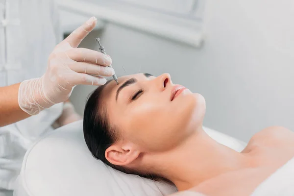 Teilansicht des Kosmetikers, der während der Akupunkturtherapie im Wellness-Salon Nadel auf die Stirn der Frau legt — Stockfoto